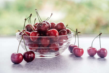 Ripe sweet cherries in a glass bowl on wooden table with green bokeh background, summer fruits
