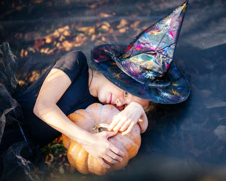 Halloween little girl in witch hat holding a pumpkin and lying on the trampoline with yellow leaves...