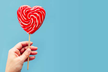 A hand holding red heart lollipop on blue background love and Valentine day backgroundの写真素材