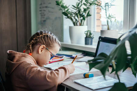 Primary school girl in eyeglasses writing in her notebook while having online lesson during ...