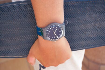 Alicante, Spain - August, 2019: Grey rubber Swatch watch on teenager's wrist holding skate boardの写真素材