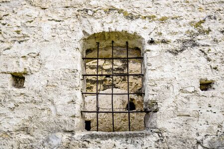 Arched window with metal bars in old stonework. The texture of the old dilapidated masonry. The ancient half-ruined synagogue. A place for religious rituals. Rashkov, Moldova. Selective focus.の写真素材