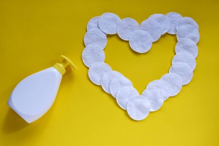 White plastic soap dispenser bottle and heart made of cotton pads on yellow background. Top view. Flat lay. Copy space for text. Selective focusの写真素材