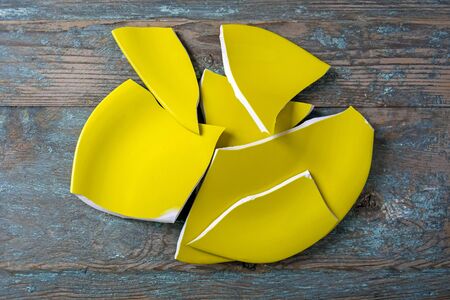 Fragments of broken yellow plate are randomly located on old wooden surface. Sharp edges. Danger of cuts. Traces of peeling paint on wooden board. Top view. Close-up. Selective focus. Copy space.の写真素材