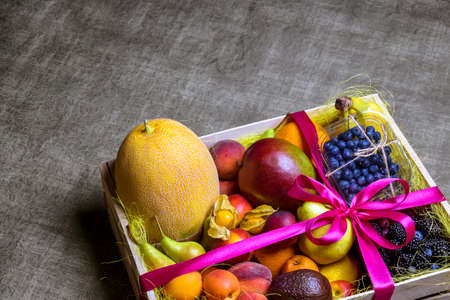 Variety of ripe fruits and berries are beautifully stacked in wooden box tied with pink ribbon. Dark background. Food delivery, gift fruit set. Concept of healthy eating. Copy space. Selective focus.の写真素材
