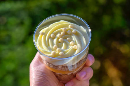 Delicious dessert in transparent glass in woman's hand against green background. Close-up. Trifle is delicate sweet dessert made of sponge dough and custard. Copy space. Selective focus.の写真素材