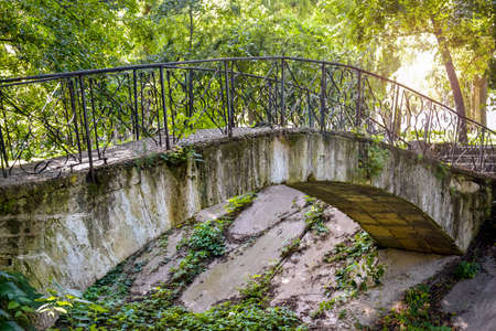 Old concrete arched footbridge with wrought iron railings against backdrop of green foliage. Dry river with concrete slabs. Ancient park in summer.の写真素材