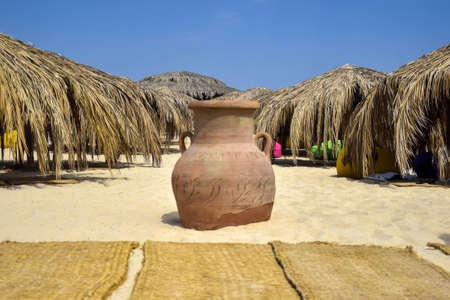 Clay urn on beach, against backdrop of umbrellas covered with dry palm leaves on bright sunny day....