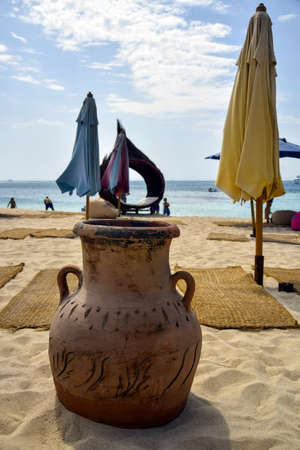 Clay urn on beach, against backdrop of folded umbrellas, sea and blue sky with fluffy clouds on bright sunny day. close-up. selective focus.の写真素材