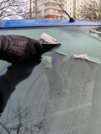 Man scrapes hoarfrost with a plastic scraper from the side window of a blue car. Hand in a black leather glove in the cold. close-up. copyspace. selective focus.の写真素材