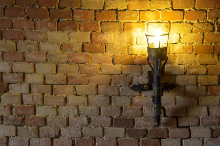 Vintage metal forged lantern hangs on old brick wall. Brightly burning lamp illuminates brickwork. Copy space for text.の写真素材