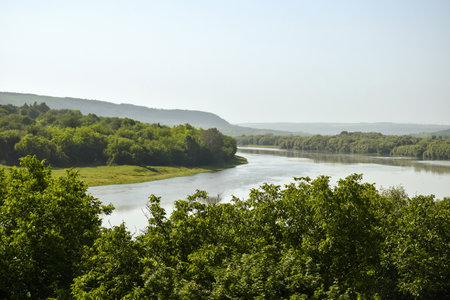 River bend with green forested banks on summer day against blue sky. Hilly terrain, slow flow.