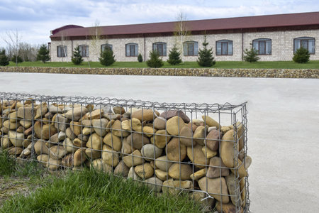 Stone fence reinforced with strong galvanized metal mesh, made of large smooth pebbles against backdrop of concrete platform and old long stone building. Copy space for text.の写真素材