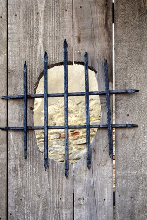 Black forged lattice on wooden round window cut into gate, darkened weathered wood. View through window to old stone dilapidated wall. Selective focus. Copy space for text.の写真素材