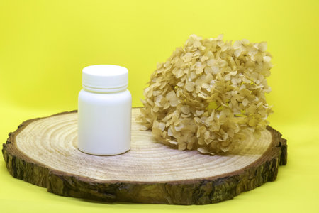 White plastic medical container with dried hydrangea flower on wooden board on yellow background, mockup. Blank Medicine bottle. Pharmacy business and treatment concept. Copy space. Close-up.の写真素材