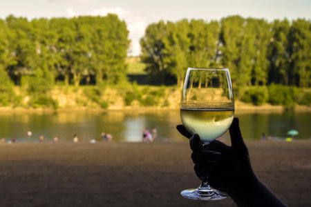 Frosted glass of cool white wine in woman hand, against blurred picturesque background of river beach with people. Close-up.の写真素材