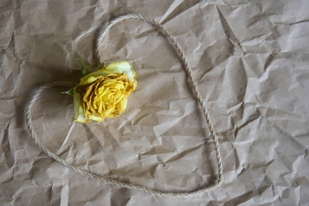Heart shape made of twine and dried yellow rose head on background of craft crumpled paper. Concept photo reflecting relationships, love and mutual understanding. Copy space. Close-up.の写真素材