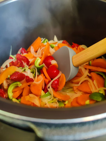 Cooking saute at home. Stewed vegetables and silicone spatula in saucepan. Mixed Ingredients onions, carrots, tomatoes, bell pepper. Close-up. Selective focus.の写真素材