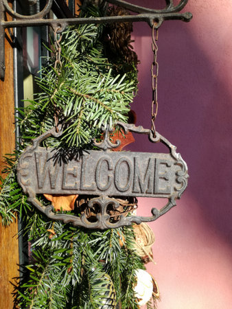 Christmas decoration. Old iron sign with inscription Welcome, decorated with coniferous garland, hangs above entrance door of old building. Close-up. Copy space.の写真素材