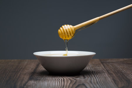 Honey dripping from a wooden honey dipper into a white bowlの写真素材