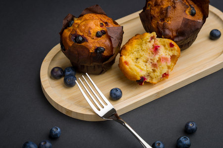 Two muffins topped with blueberries sit on a wooden tray. A fork is next to the muffins, and fresh blueberries are scattered on the surface. One muffin is partially eaten.の写真素材