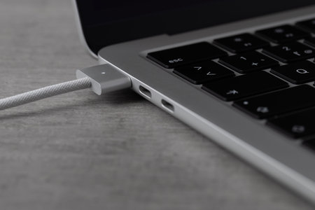 A MacBook is connected to a charging cable through its USB port. The device sits on a wooden table,...
