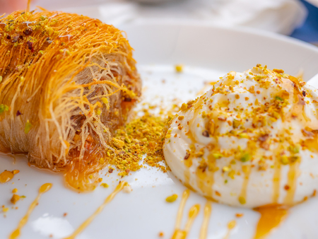 Close up of a succulent Greek dessert. Greek kataifi with honey yogurt and grated pistachios on a white plate and a blurred background. Horizontal viewの写真素材