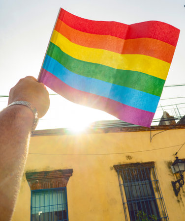 A hand holds a rainbow flag of the LGBTQ movement, house in backgroundの写真素材
