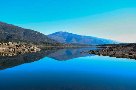 Trunk of an old tree on the shore of a lake with calm water and reflection of the mountains in the...