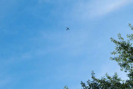 A propeller plane flies in a blue sky with few clouds in the background and tree branches on one...