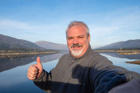 Mature hiker taking a selfie portrait on top of the mountain next to a lake.