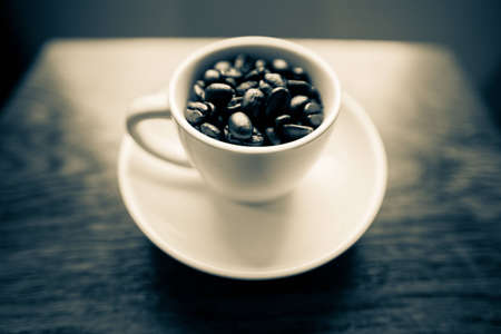 A black and white shot with a bit of a blue hue of a white coffee mug on a small white plate filled with dark roasted coffee beans.  The mug is on a cherry wood table.の写真素材