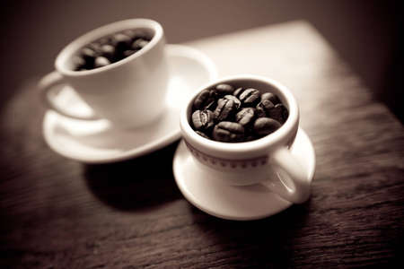 Two white coffee mugs on small white plates filled with dark roasted coffee beans.  The mugs are on a cherry wood table.  の写真素材