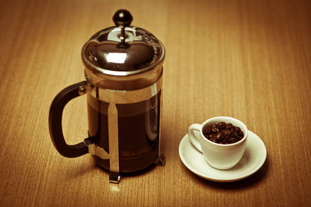A white coffee mug on a small white plate filled with dark roasted coffee beans sitting next to a french press. の写真素材