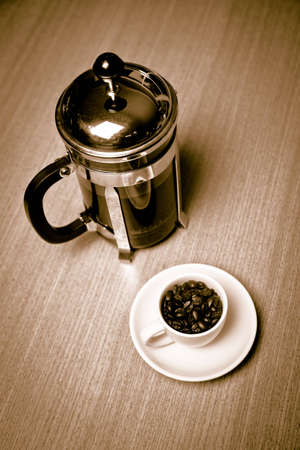 A french press sits next to a white coffee mug on a small white plate filled with dark roasted coffee beans.  The mug is on a cherry wood table.の写真素材