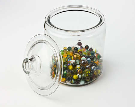 Glass jar half full of colorful glass marbles on a white background. There are a variety of different colors including yellow, green, orange, blue, black, and red.の写真素材