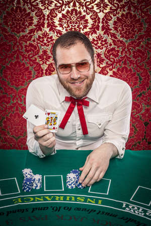 A man wearing glasses, a white shirt, and a red Texas tie sits at a blackjack table  He holds up a winning hand and smiles at the camera  There are stacks of blue and white chips in front of him のeditorial素材