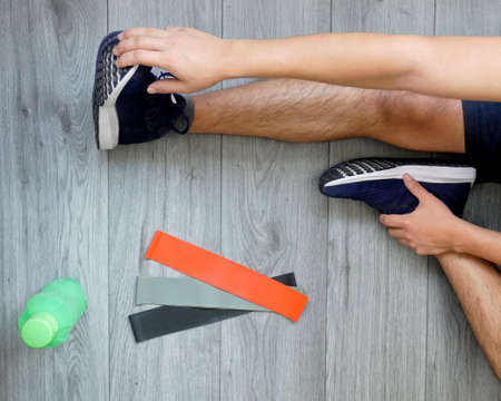 young man stretching legs at home at the end of a routine, wearing blue tennis shoes on a gray wooden floorの写真素材