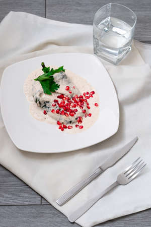 Chile en Nogada, Mexican dish that dates back to the colony, poblano pepper with a cream of walnut, with grains of pomegranate, in a white plate that is on a gray wooden table, next to it is a napkin, fork, knife and a glass of waterの写真素材