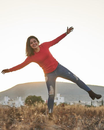 Woman in blue jeans, red sweater, in profile watching the sunrise in a golden field and the sun in...