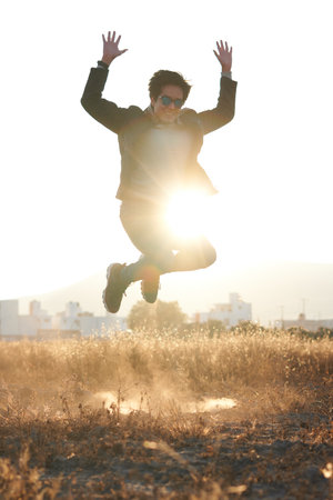 Man jumping in jeans, blue jacket, white t-shirt, with glasses, in profile looking at the sunrise in a golden field and the sun in the background peeking from a mountainの写真素材