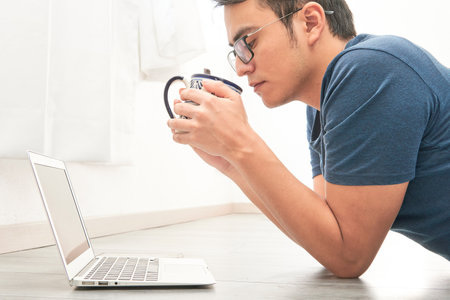 Young man having a coffee in front of the computer reading the latest news, watching a video lying...