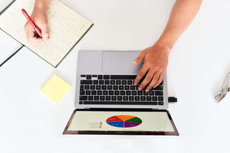 latin man typing on laptop computer on his white desk and taking notes in his diary with a red pen, top view, shot from above, model not recognizable.の写真素材