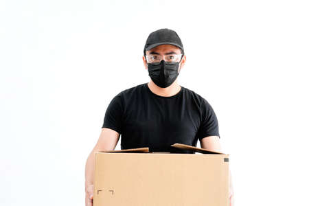 Young male delivery man, wearing health mask, glasses, protecting it from pandemic, dressed in black, with cap, holding a box, on white background, Latino.の写真素材