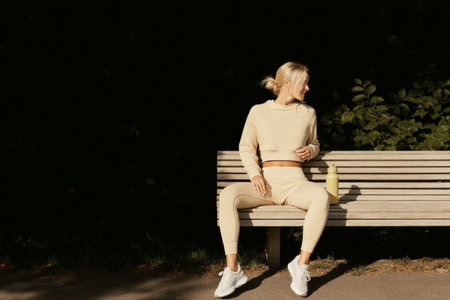 Blonde caucasian female in activewear sitting on park bench with drink bottle.の素材