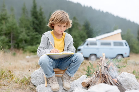 Young caucasian boy sketching by campfire with camper van in forested mountain landscape.の素材