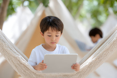 Asian child on hammock using tablet in outdoor setting.の素材