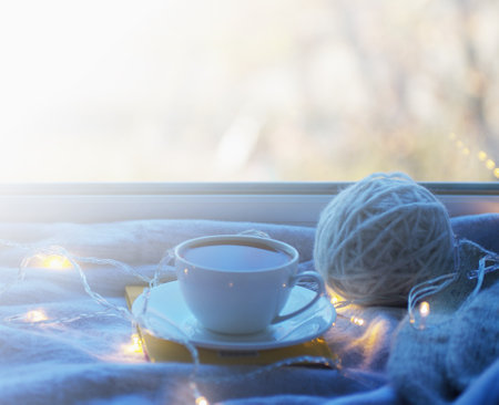 Cozy winter still life: mug of hot tea and book with warm plaid on windowsill against snow landscape from outside.の写真素材