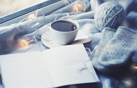 Cozy winter still life: mug of hot tea and book with warm plaid on windowsill against snow landscape from outside.の写真素材