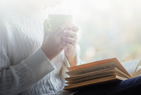 Girl is holding a cup of tea and reading a book beside the windowの写真素材